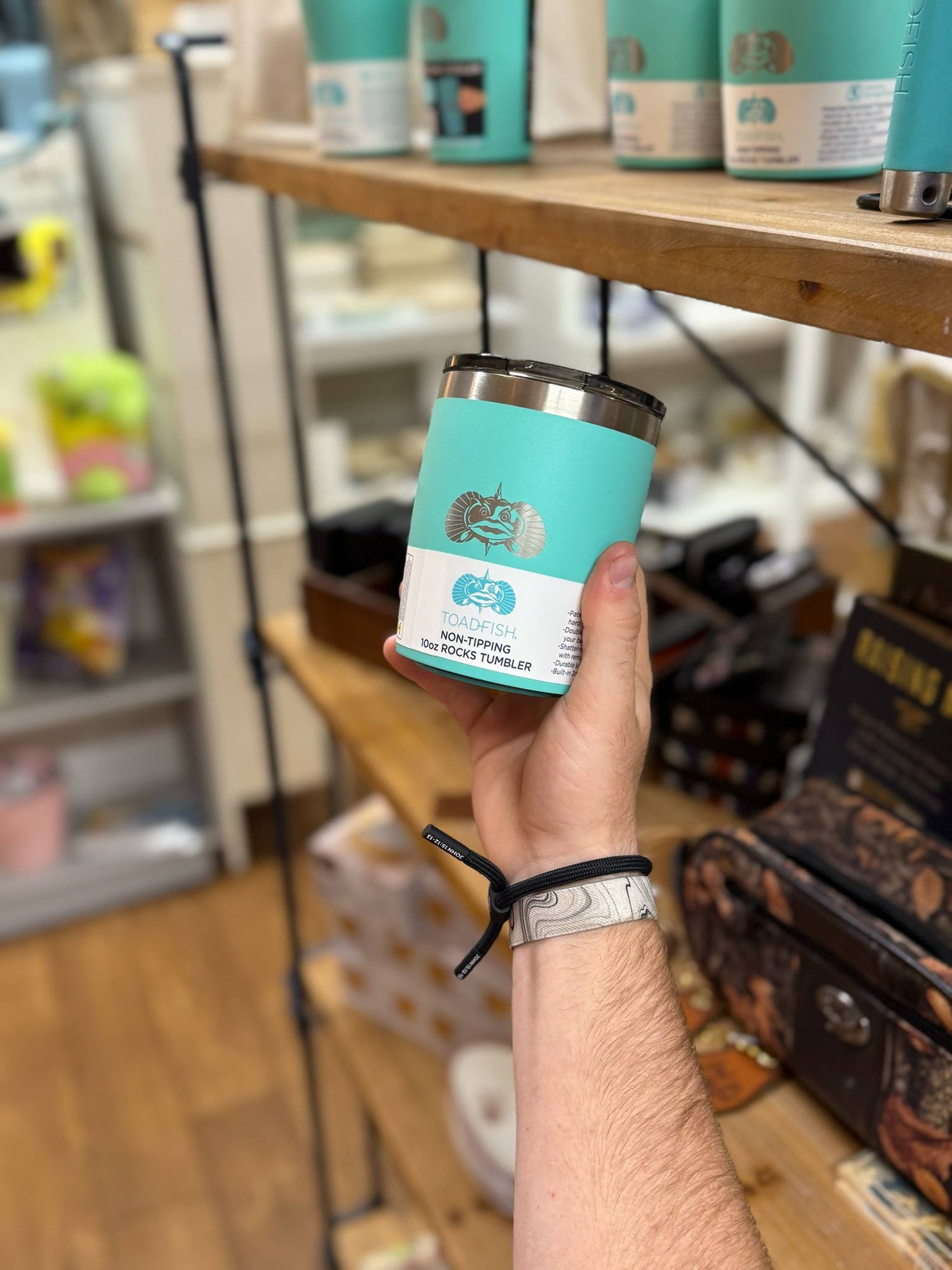 Close-up of a hand holding a Toadfish 10oz non-tipping rocks tumbler in teal, displayed on a wooden shelf at Shoppes on Davis.
